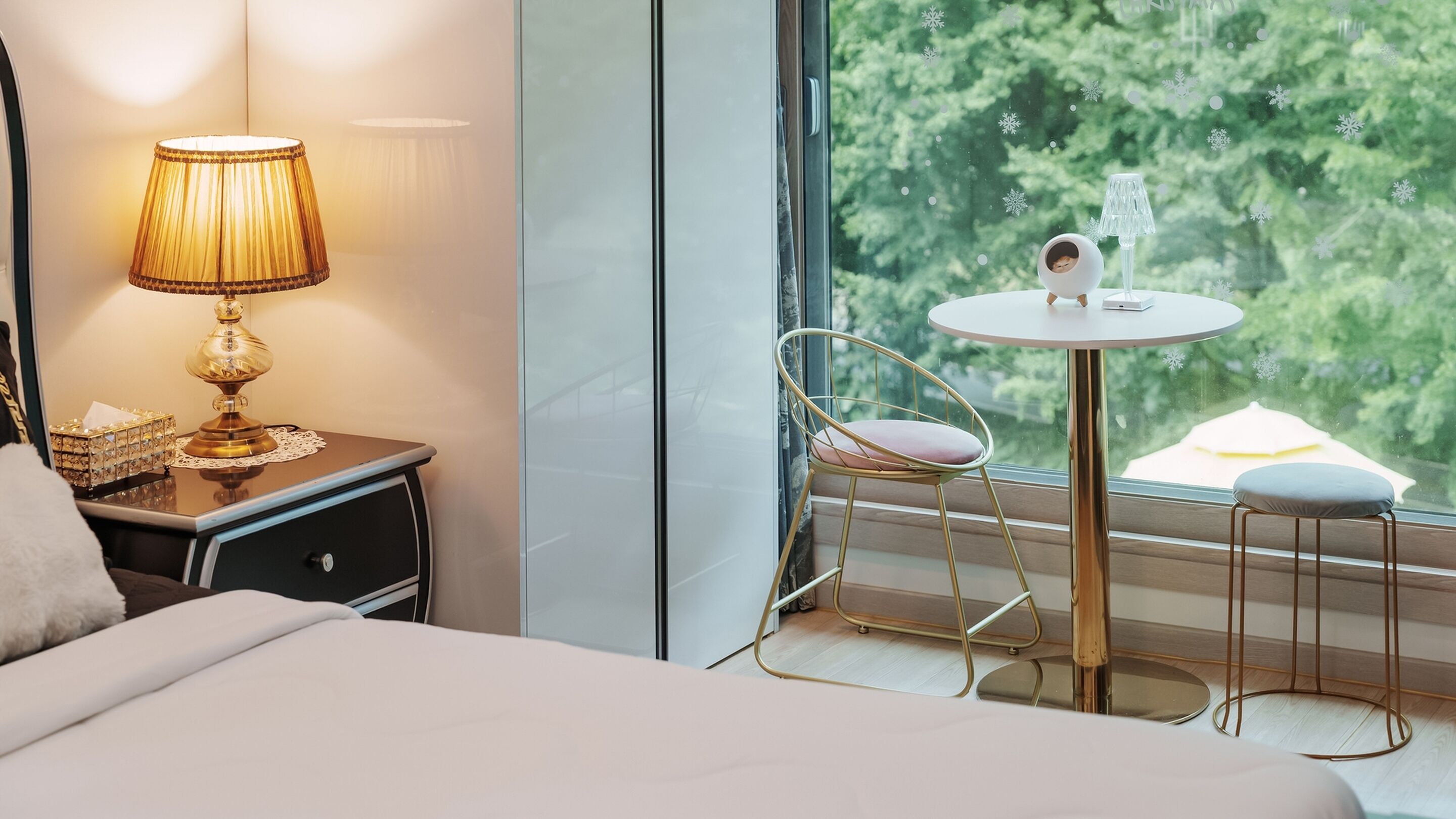 A hotel room with a bed, a nightstand with a lamp, a small table with a chair, and a view of trees through the window.