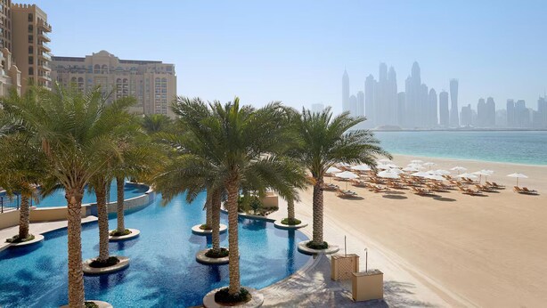 A resort pool with palm trees, a sandy beach, and a city skyline in the background.