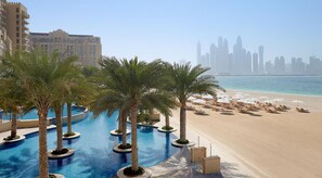 A resort pool with palm trees, a sandy beach, and a city skyline in the background. - Fairmont North Residences -Palm Jumeirah (Dubai)