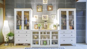 A room with a central console table, flanked by two white cabinets with glass doors. The cabinets contain books and decorative items. Above the table, there are framed pictures on the wall. - From H Amoremio The Classic Suwon (Suwon)