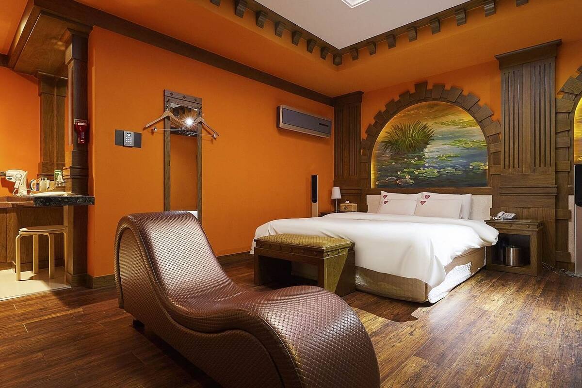 A hotel room with a bed, a chair, a desk with a coffee maker, and a painting of a landscape.