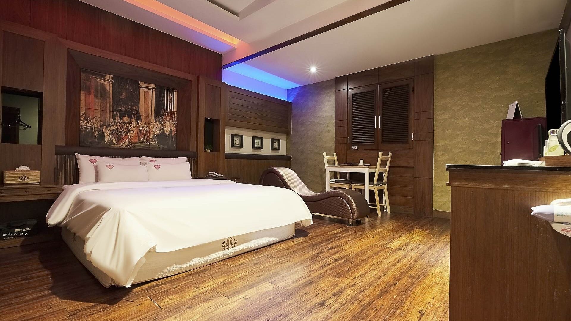A hotel room with a large bed, a desk, and a chair. There is a painting on the wall and a mirror above the bed.
