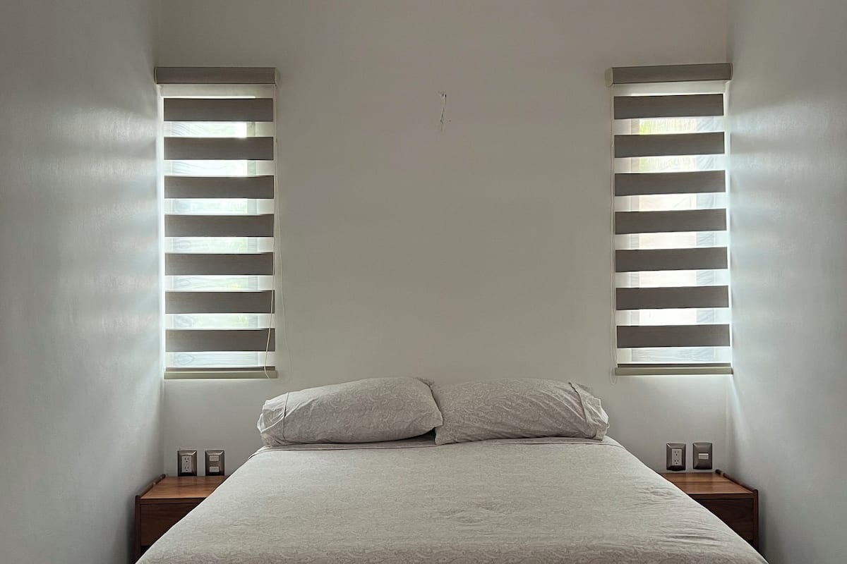 A bedroom with a bed, two nightstands, a ceiling fan, and two windows with blinds.
