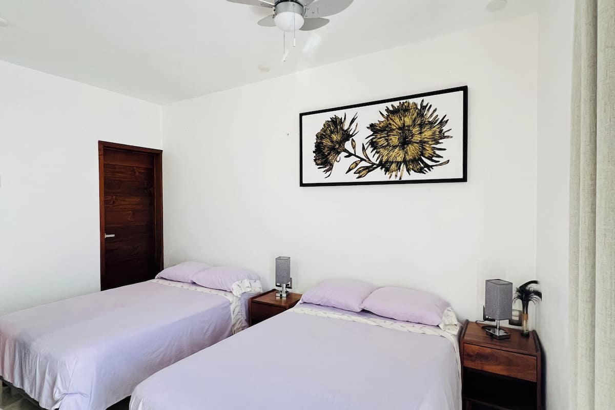 A bedroom with two beds, a ceiling fan, a framed artwork, bedside tables, and a door.