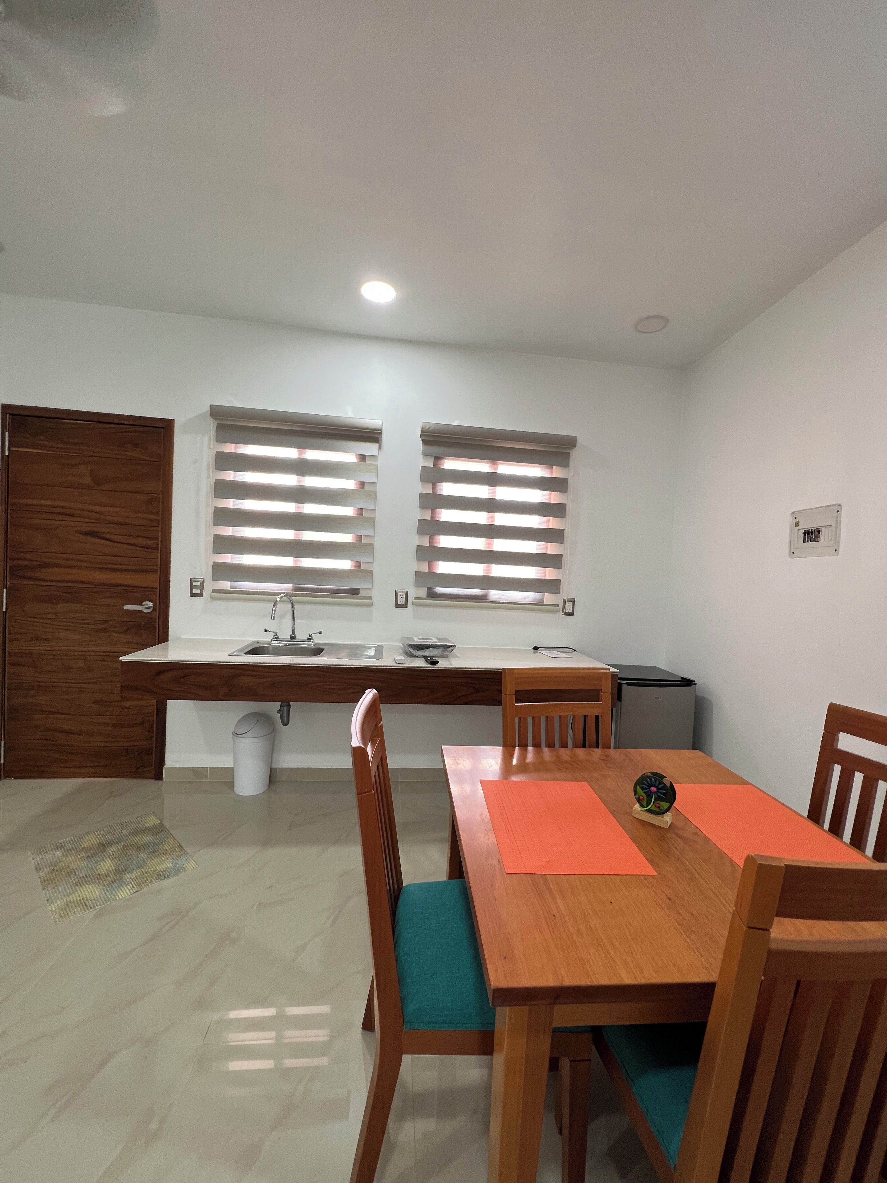 Cozinha compacta com pia, duas janelas com persianas, mesa de madeira com cadeiras e lixeira.