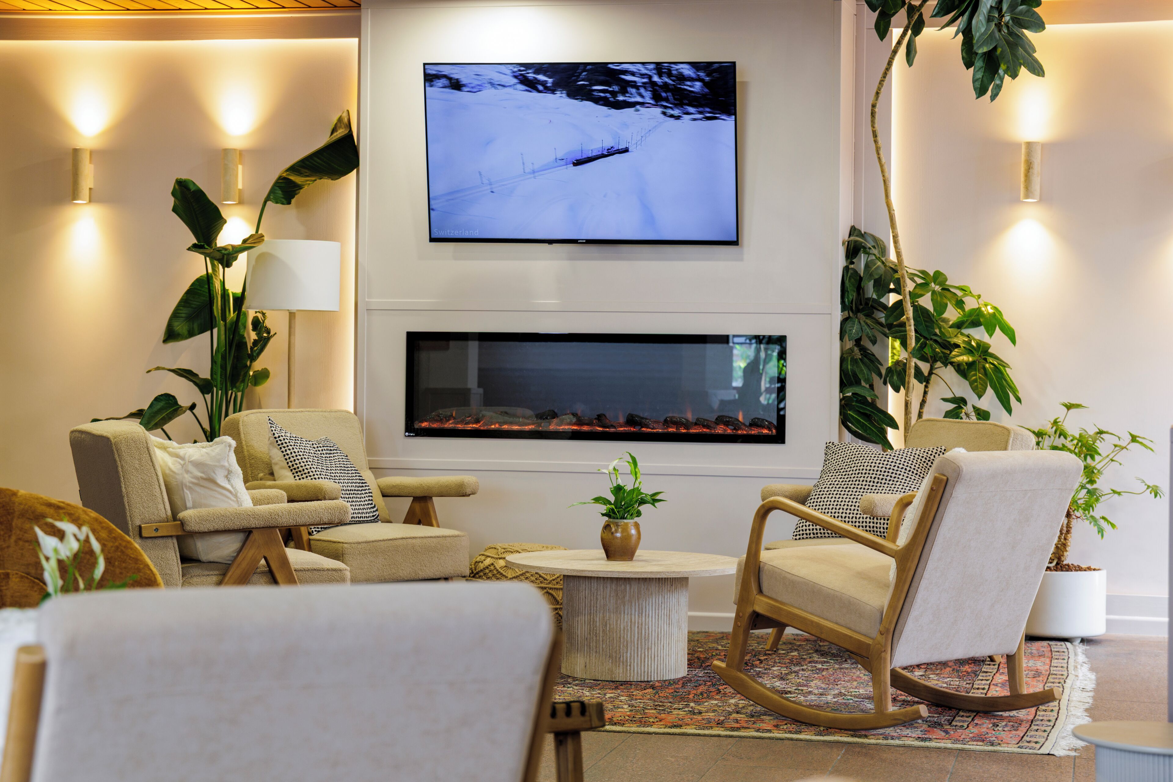 Uma sala de estar moderna com uma TV de tela plana na parede, lareira abaixo, assentos confortáveis e plantas decorativas.