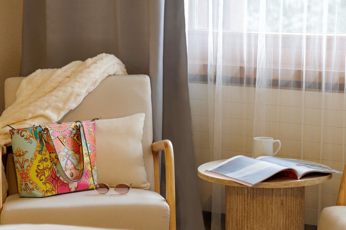 A hotel room with a chair, a table with a book, a pair of glasses, and a colorful bag.
