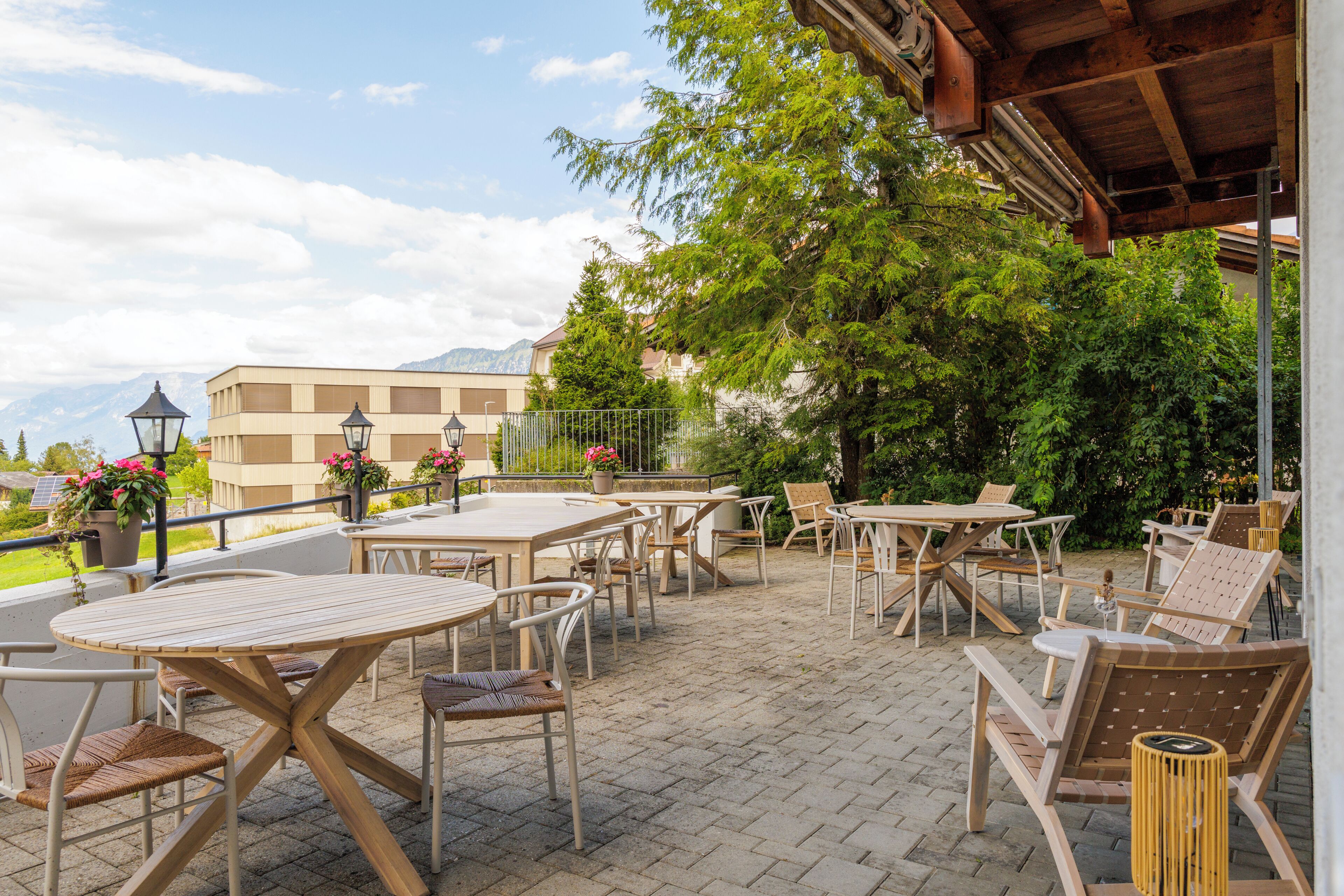 An outdoor patio with tables and chairs, surrounded by greenery and a view of mountains.