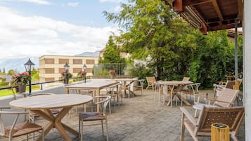 An outdoor patio with tables and chairs, surrounded by greenery and a view of mountains.