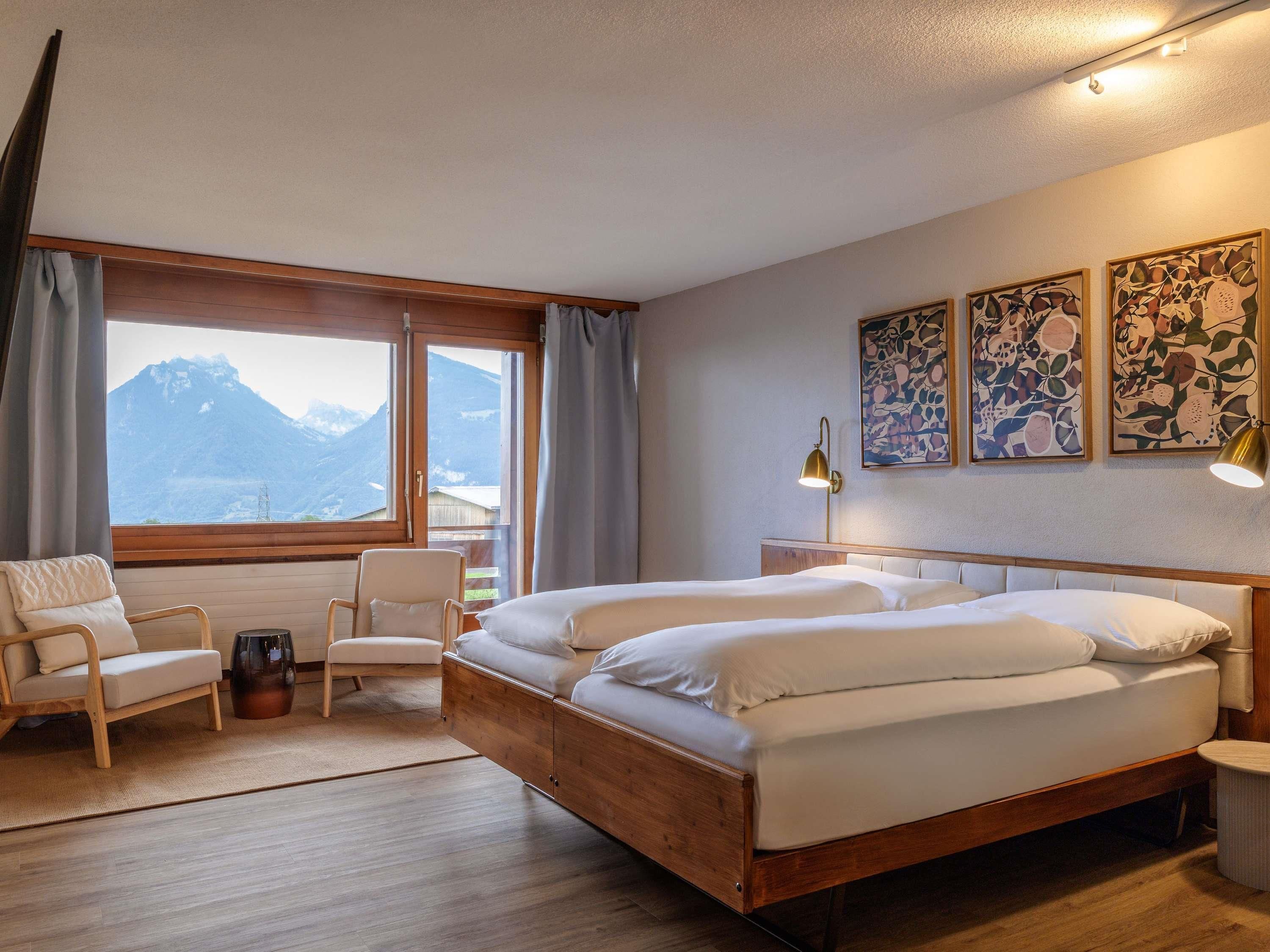 A hotel room with a large bed, two chairs, a bedside table, and a view of mountains through a large window.
