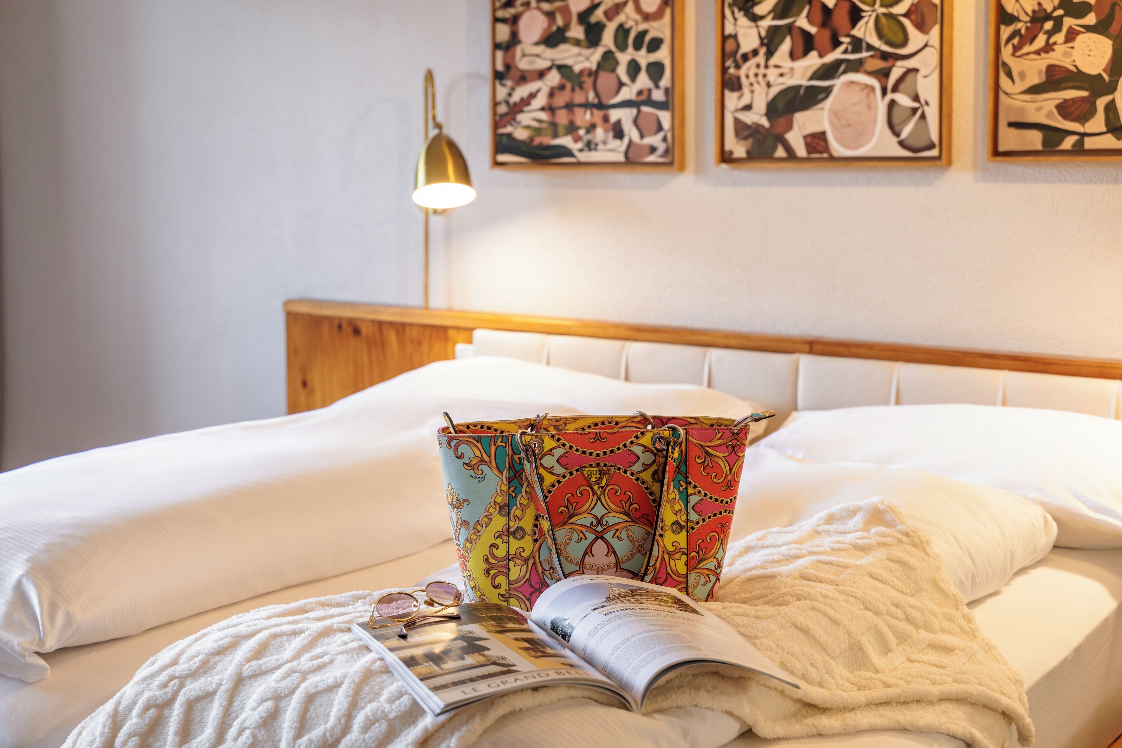 Un lit bien fait, agrémenté d’un coussin coloré, d’un magazine et d’une paire de lunettes posés sur le lit.