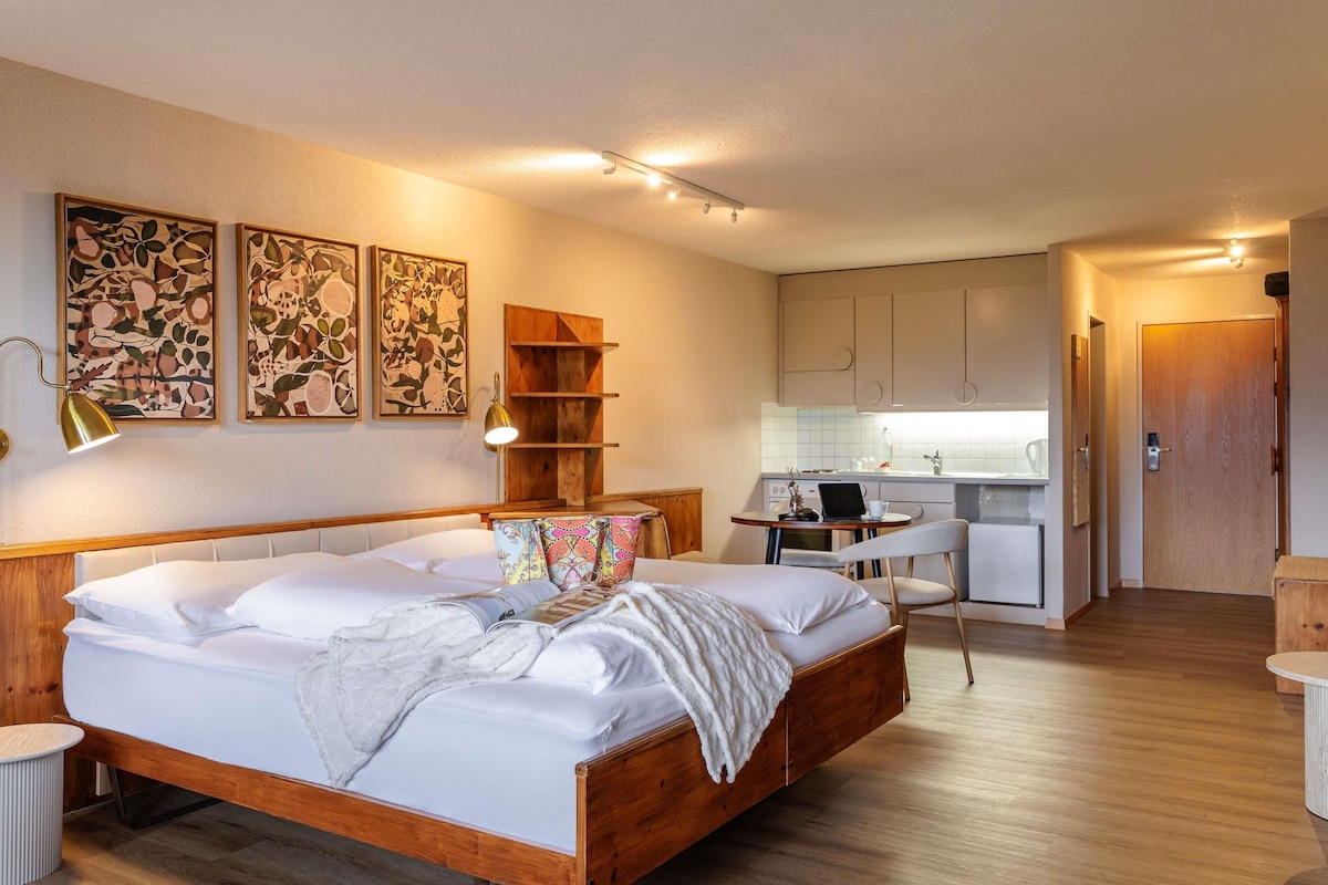 A modern bedroom with a wooden bed, bedside lamps, and a small table. A kitchenette with a sink and a chair is visible in the background.