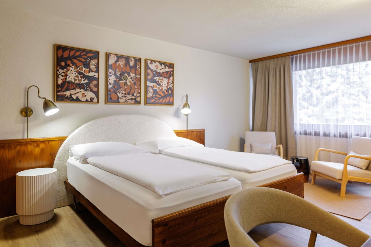 A hotel room with a double bed, two bedside lamps, a chair, a desk, and a window with curtains.