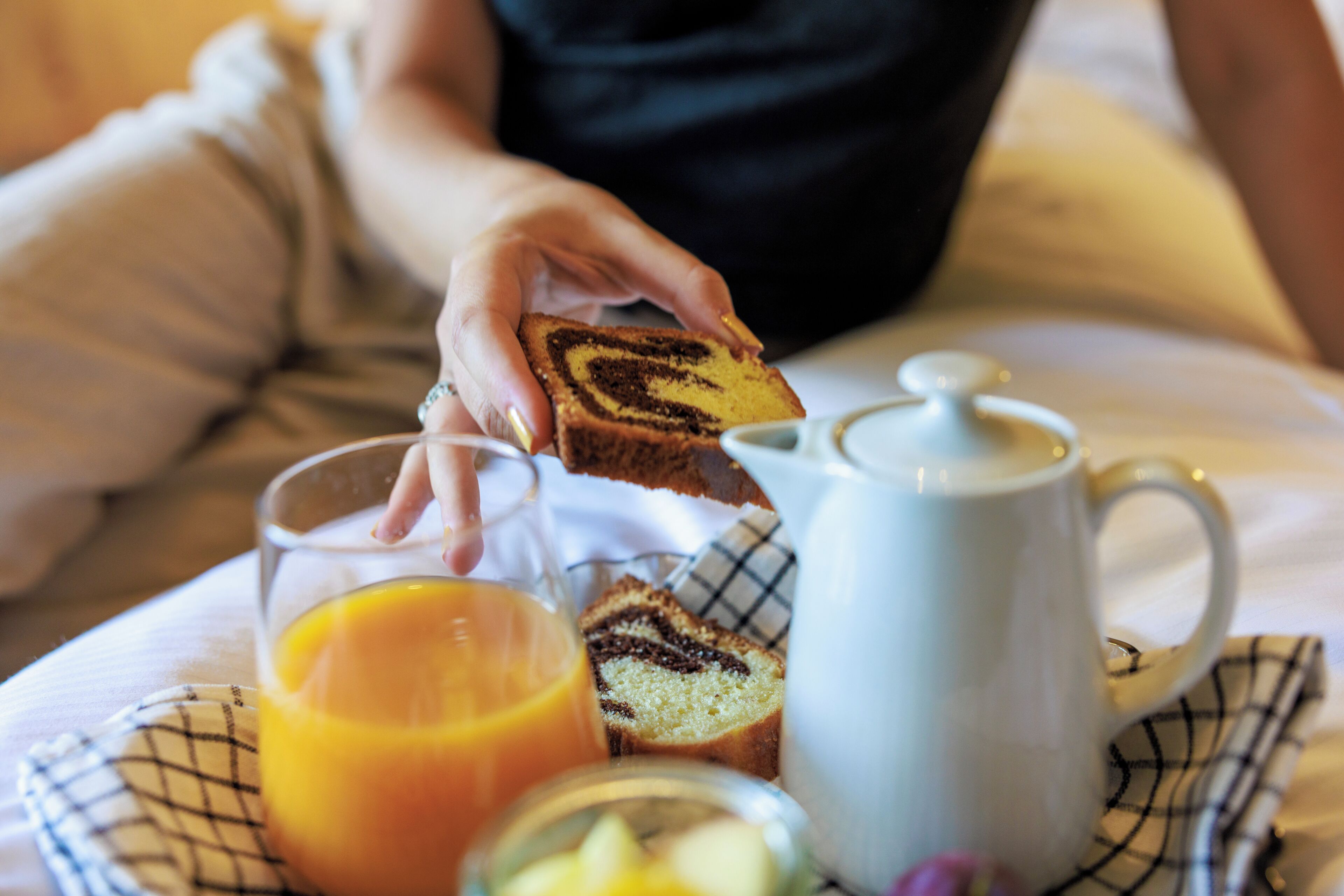 A breakfast tray with a slice of cake, a glass of orange juice, and a teapot on a checkered cloth.