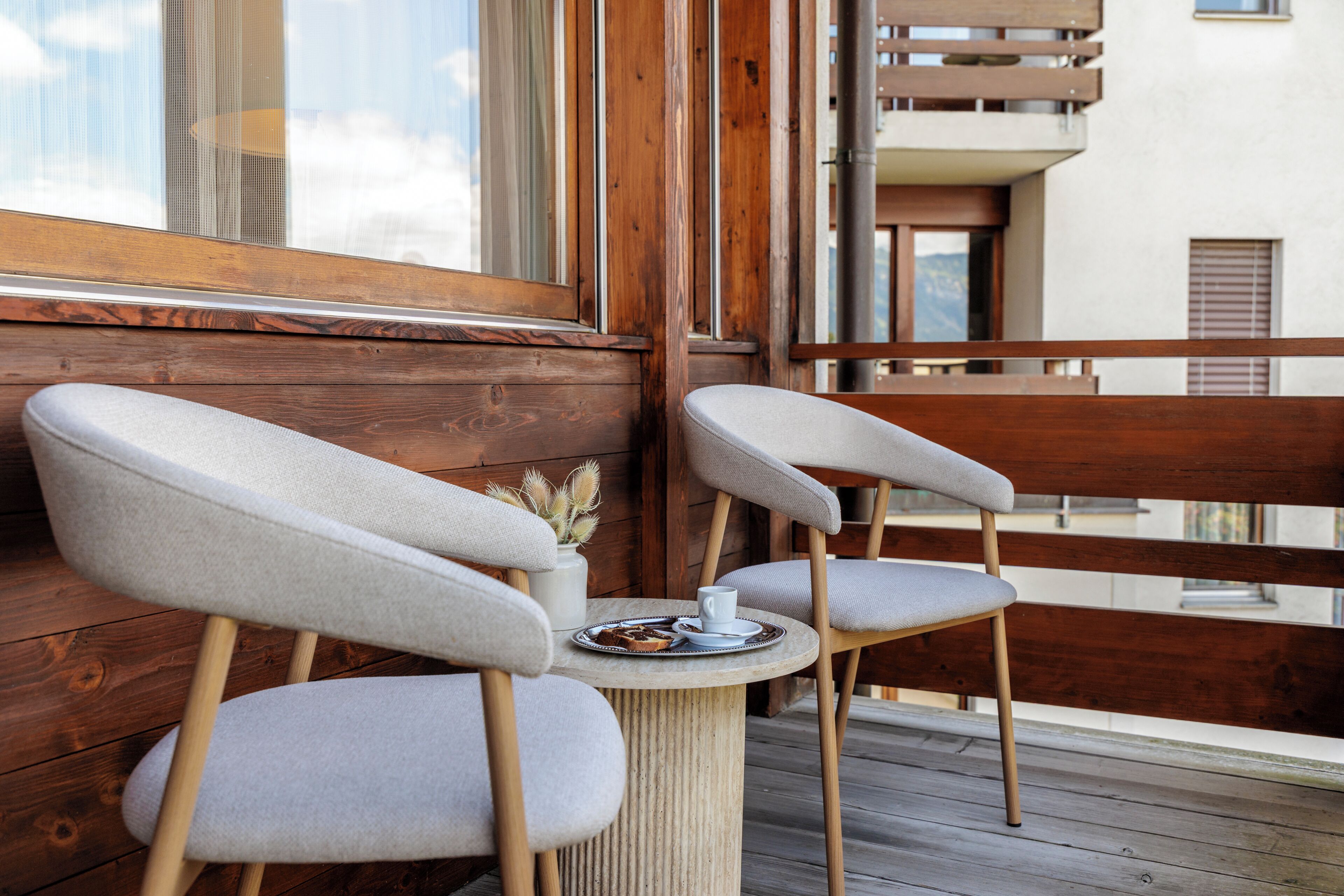 Deux fauteuils rembourrés, une petite table et une tasse sont placés sur un balcon doté de balustrades en bois.