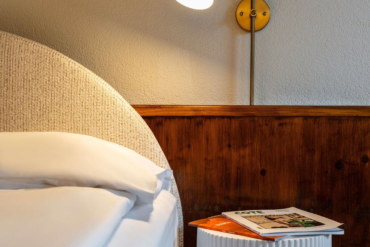 A hotel room with a bed, a wooden headboard, a wall-mounted light fixture, and a small table with a book.