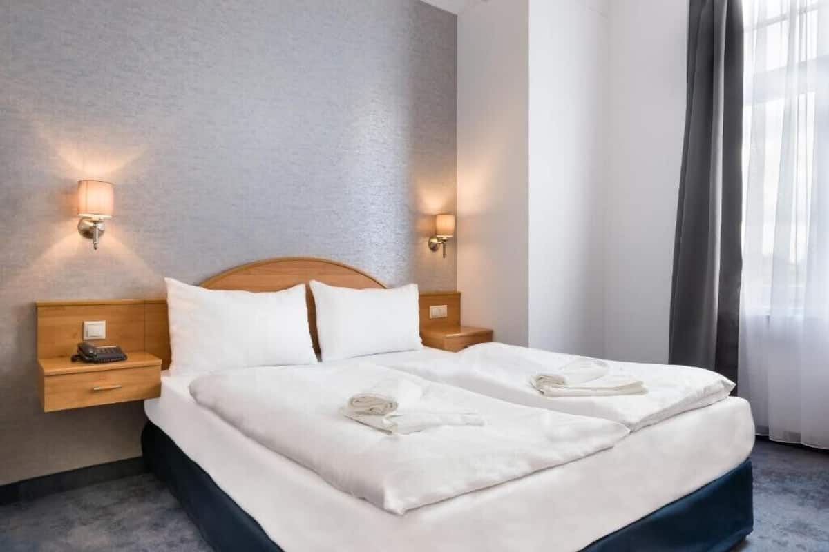 A double bed with white linens and two towels, bedside tables with lamps, and a textured grey wall.