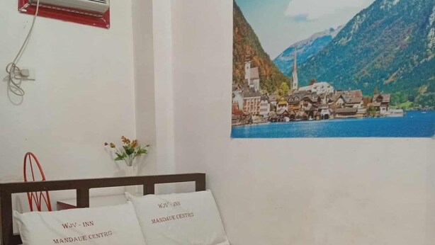 A bed with white bedding and pillows, a red-framed air conditioning unit, and a framed picture of a coastal village on the wall. - WJV INN MANDAUE CENTRO (Mandaue)