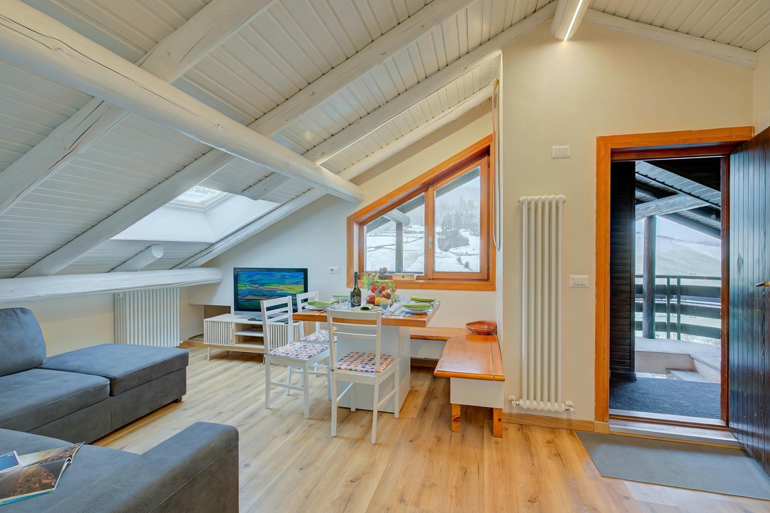 A modern loft bedroom with a sloped ceiling, skylight, wooden flooring, a dining area with a table and chairs, and a view of a snowy landscape through a window.