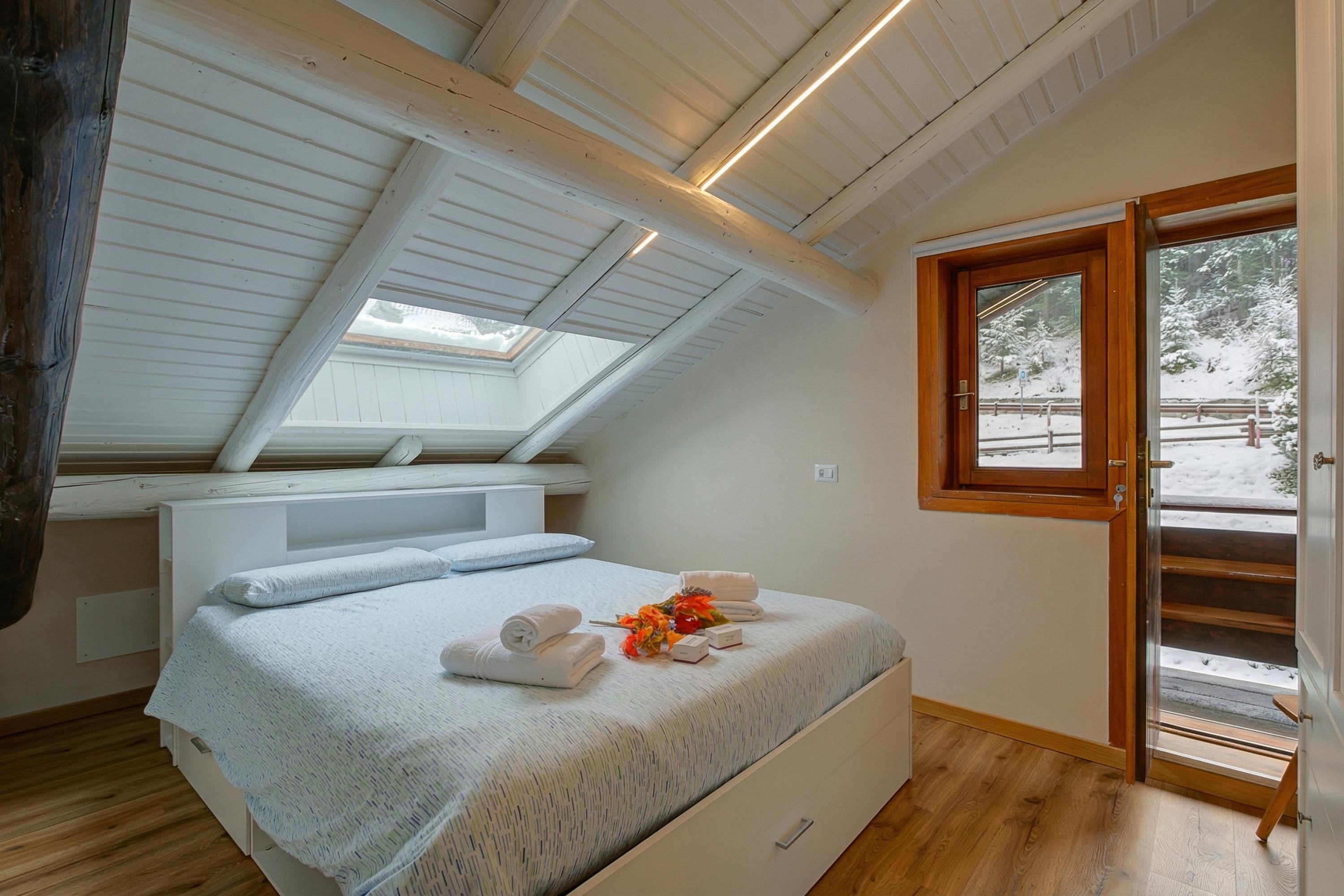 A bedroom with a sloped ceiling, a skylight, a bed with towels and a flower arrangement, and a view of a snowy landscape through a door.