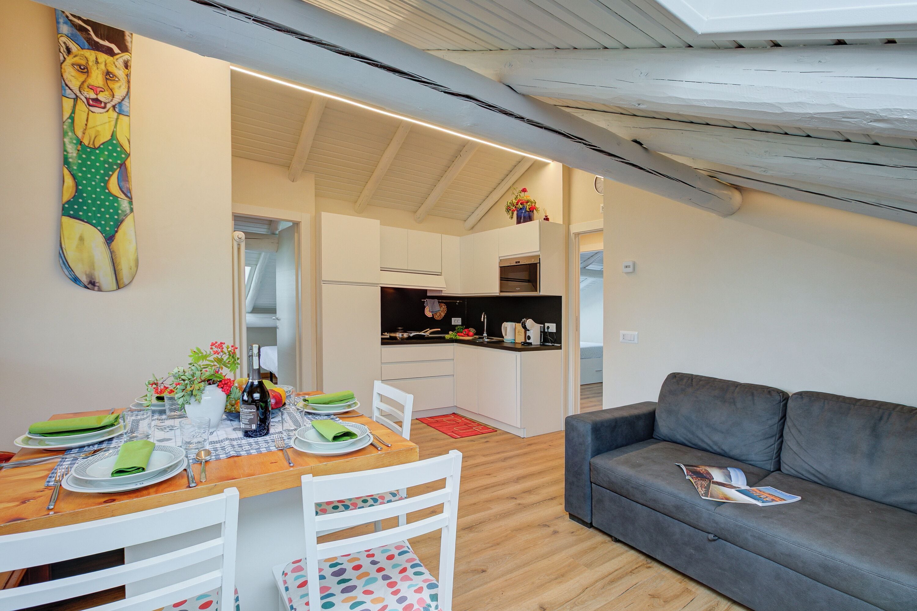 A modern living room with a grey sofa, a dining area with a wooden table set for two, and a kitchen in the background.