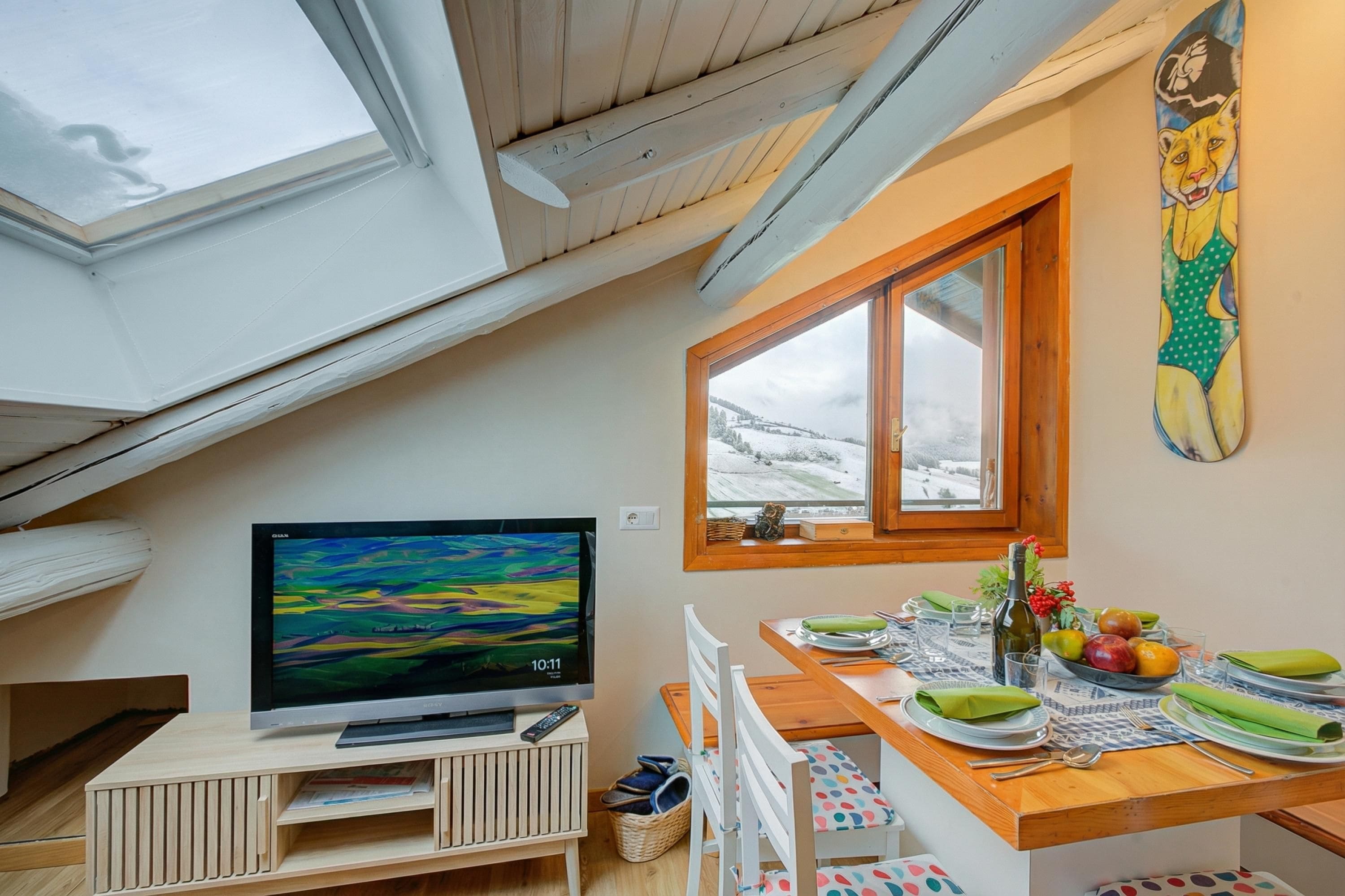A cozy room with a TV on a wooden stand, a dining table set with plates and glasses, and a window with a view of a snowy landscape.