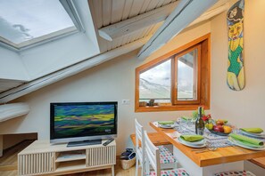 Un coin repas avec une table dressée pour deux personnes, un téléviseur posé sur un meuble et une fenêtre donnant sur l’extérieur.