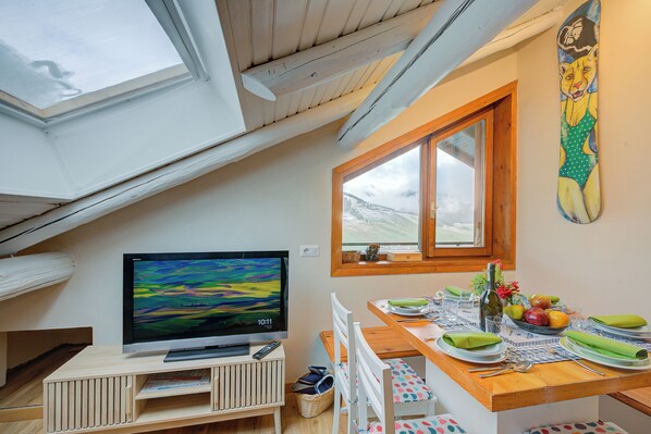 A dining area with a table set for two, a television on a stand, and a window with a view of the outdoors.