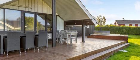 A modern outdoor patio with a wooden deck, outdoor seating, and a covered area with large windows.