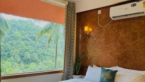 A hotel room with a large bed, a ceiling fan, an air conditioning unit, a window with a view of a forest, and a wall-mounted lamp. - Vayalada Garden resort (Koyilandy)