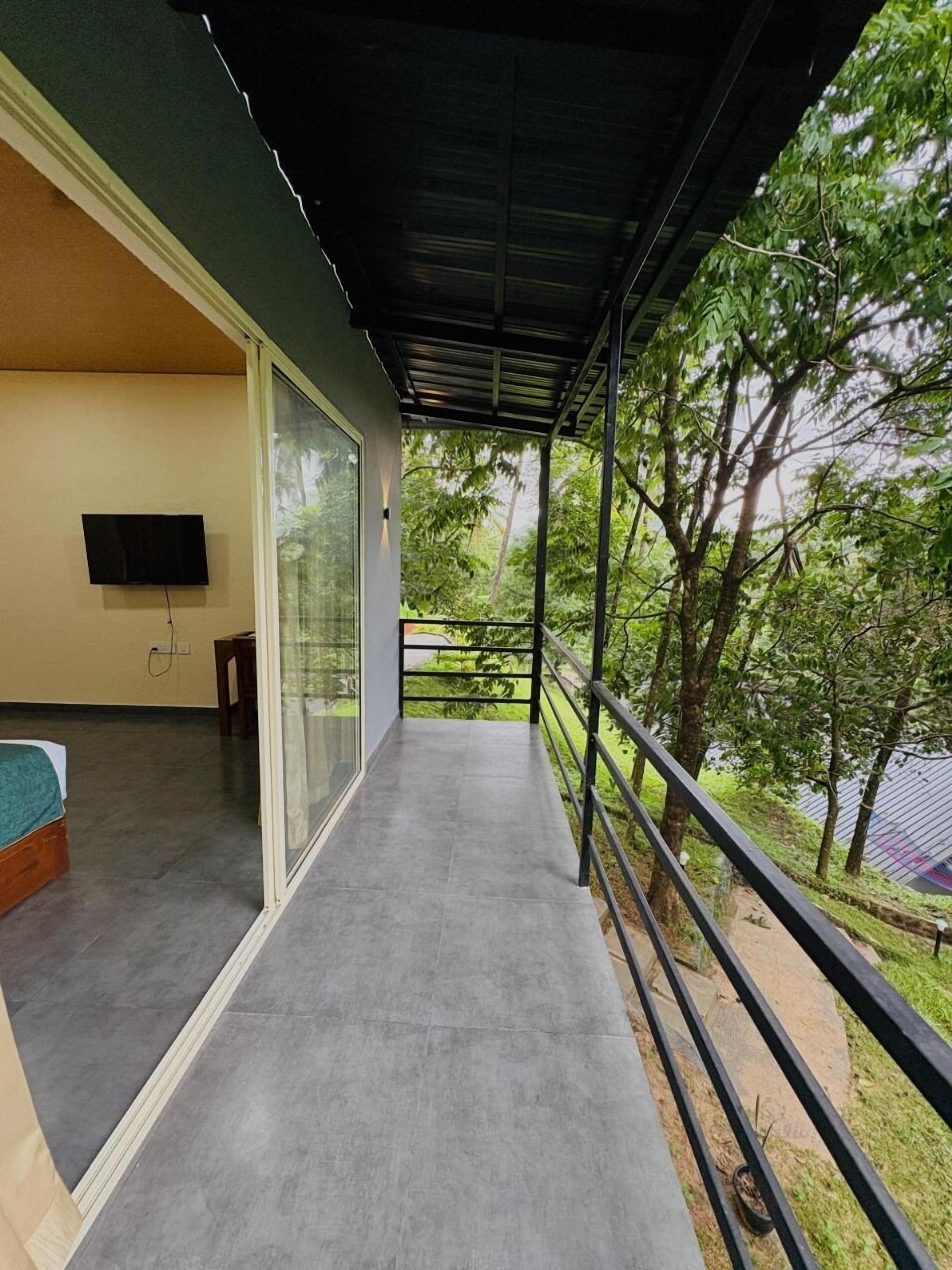 A balcony with a glass door leading to a room, a TV mounted on the wall, and a view of greenery outside.