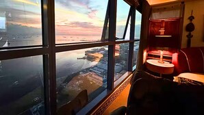 A room with a large window offering a view of a cityscape and water at sunset. There is a small table with a lamp and a chair. - Sea Family Room in Front of US Embassy (Manila)