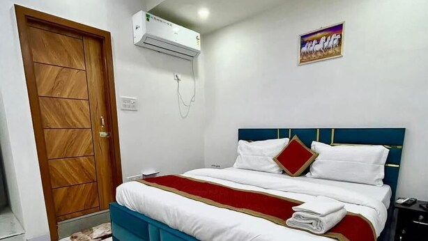 A bedroom with a bed, a wooden door, an air conditioning unit, and a wall-mounted artwork. - Roomshala Hotel J JOy (New Delhi)