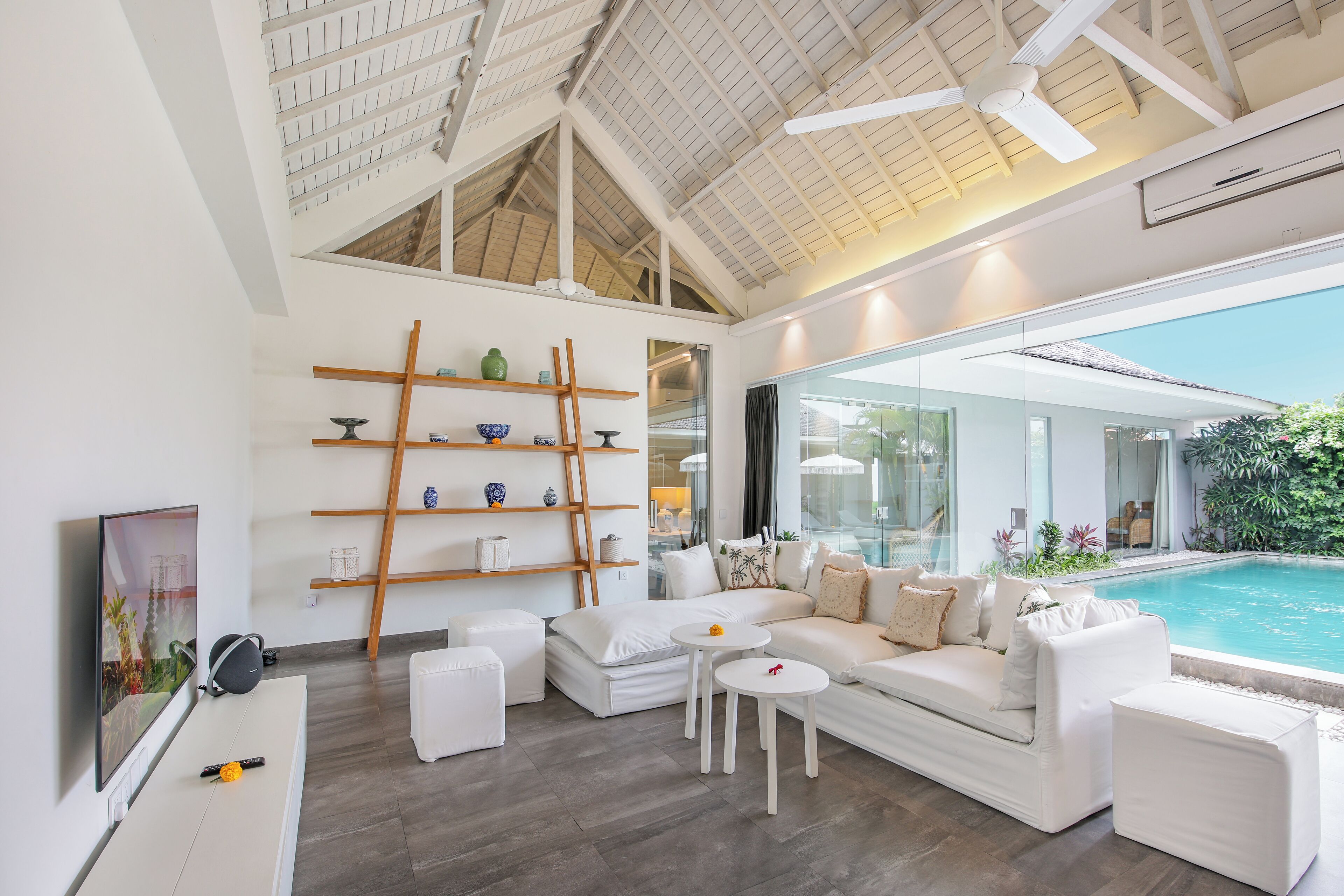 A modern living room with a white sofa, a coffee table, and a TV. There is a pool visible through the glass wall.