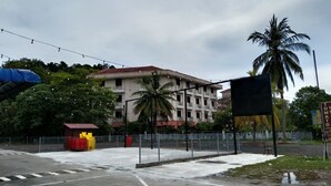 A parking area with marked spaces, a fenced playground, and a building with multiple floors in the background. - Tomato Motel (Langkawi)