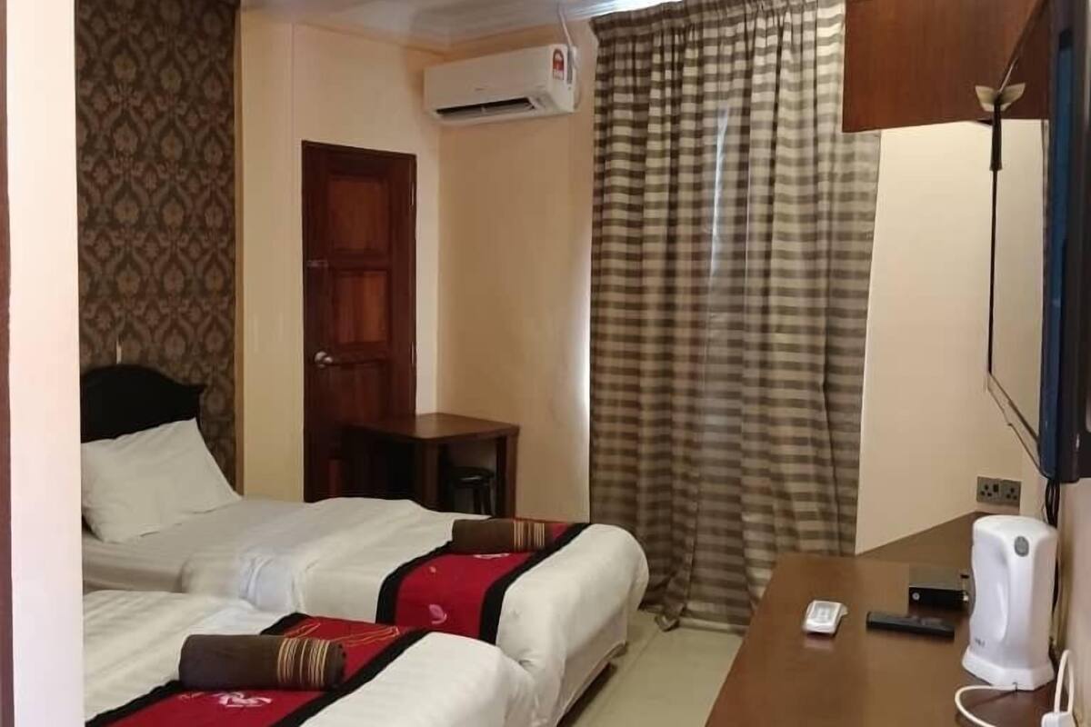 A hotel room with two beds, a desk with a phone and a coffee maker, an air conditioning unit, and a window with curtains.