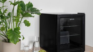 Un refrigerador negro con puerta de cristal, una planta en maceta, dos vasos, un control remoto y un cuenco de uvas sobre una superficie de madera.