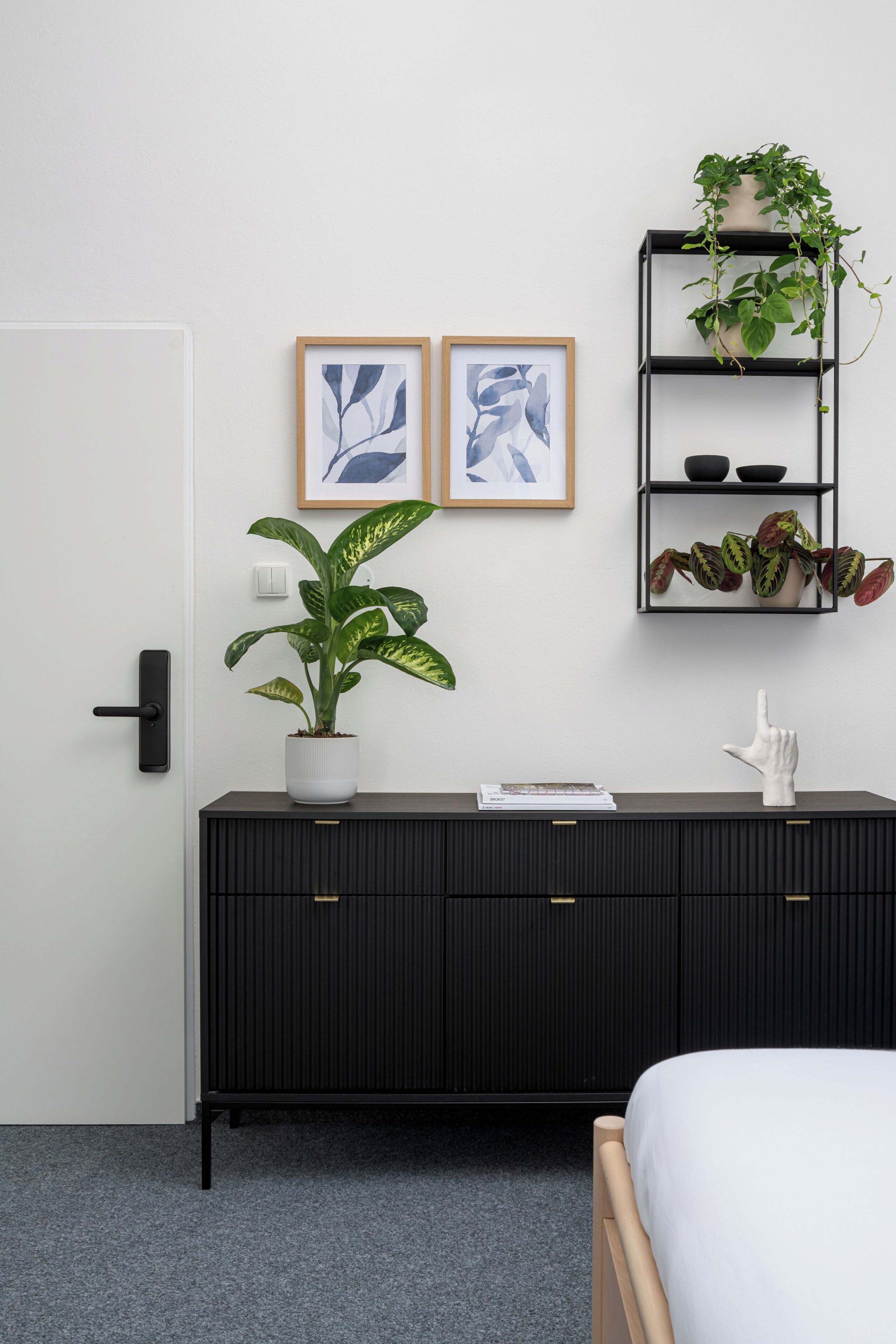 A bedroom with a bed, a black cabinet with drawers, a potted plant, and two framed artworks on the wall.