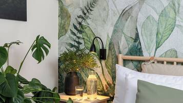 Une chambre à coucher comprenant un lit, une table de chevet, une lampe, une plante en pot et un mur orné d’un motif de feuilles.