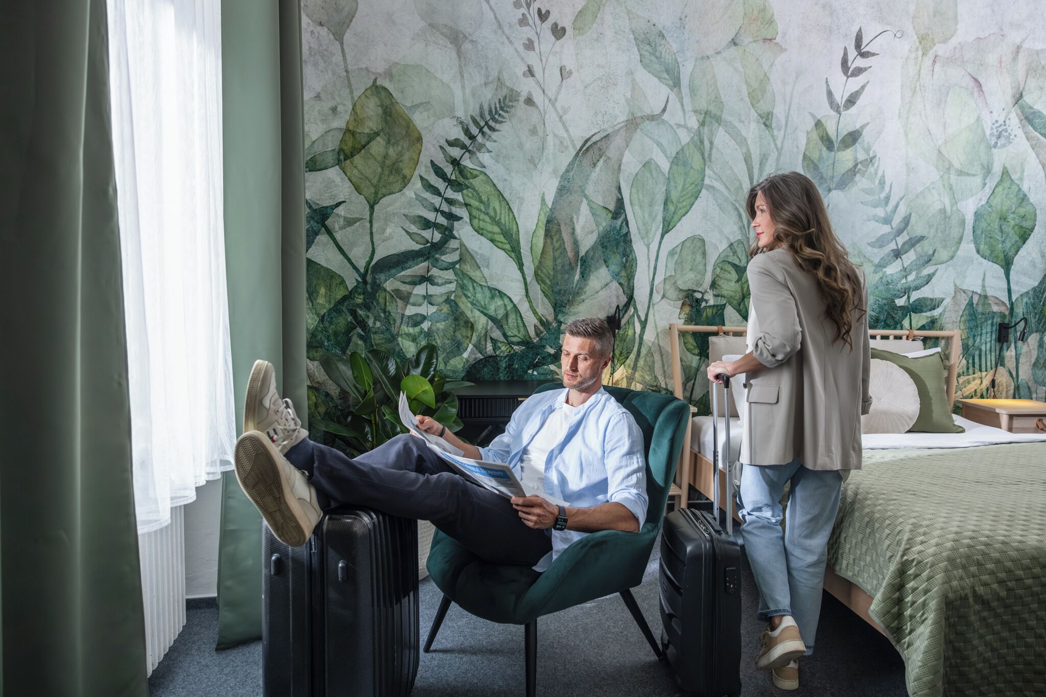 Een kamer met een bed, een groene fauteuil, een koffer en een persoon die een tijdschrift leest.