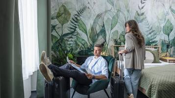 Een kamer met een bed, een groene fauteuil, een koffer en een persoon die een tijdschrift leest.