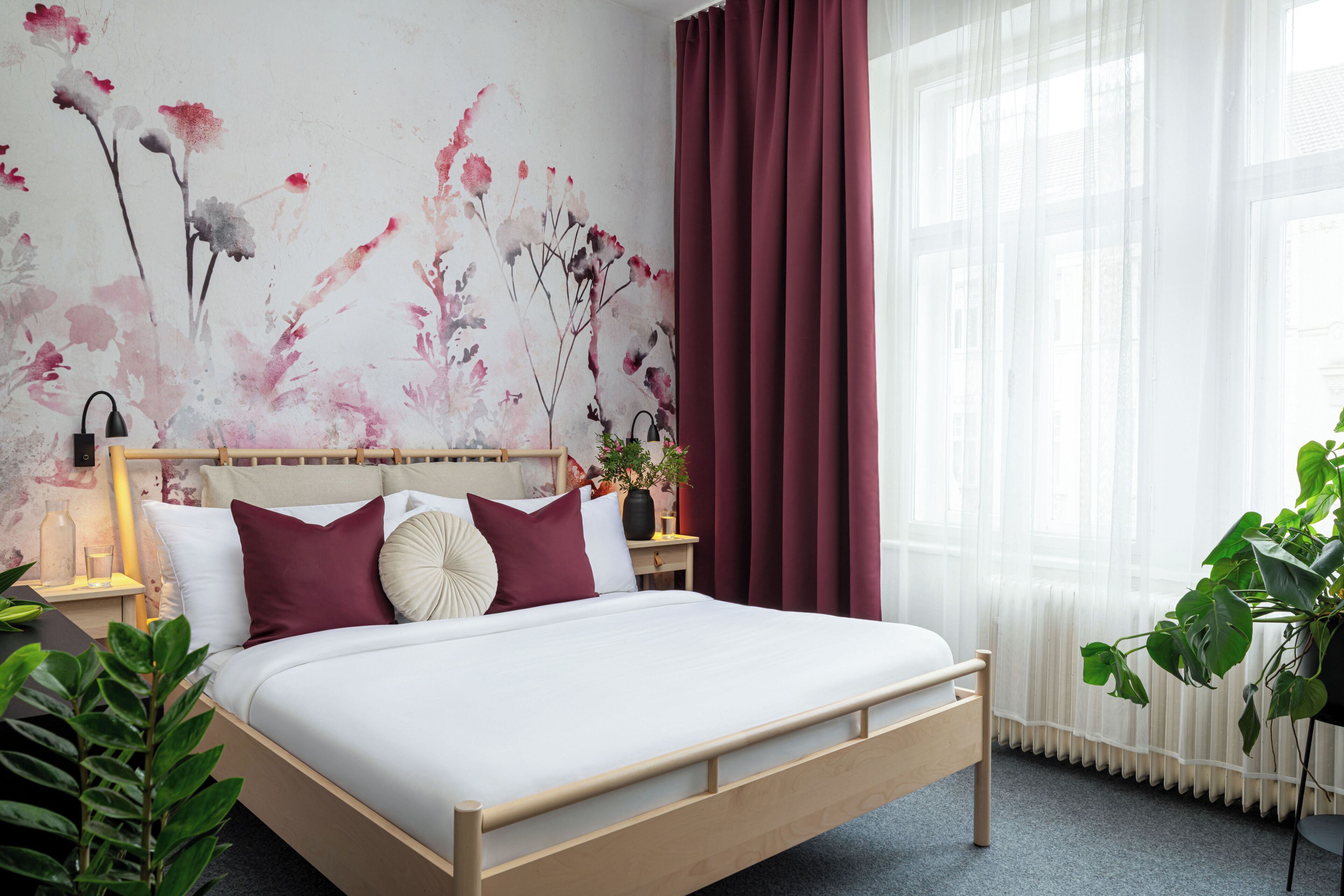 A bedroom with a wooden bed, a nightstand with a lamp, a wall with floral artwork, and a window with curtains.