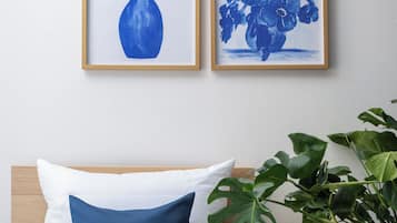 Un lit avec une literie blanche et bleue, une plante posée sur une table de chevet et deux tableaux représentant des motifs floraux, encadrés, accrochés au mur.
