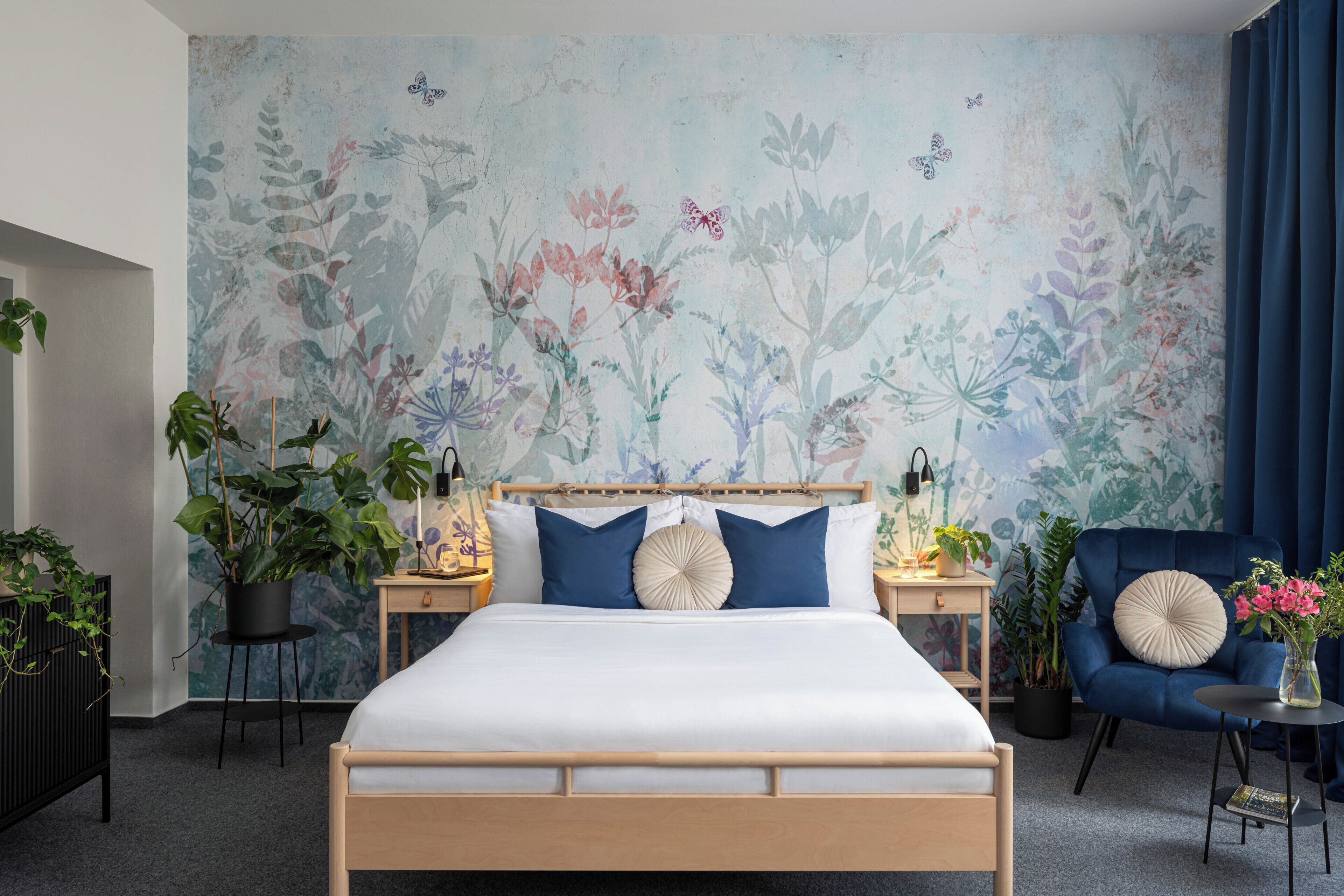 Ein Schlafzimmer mit einem großen Bett, zwei Nachttischen, einem Beistelltisch, einem blauen Sessel und einer Wand mit floralem Wandbild.