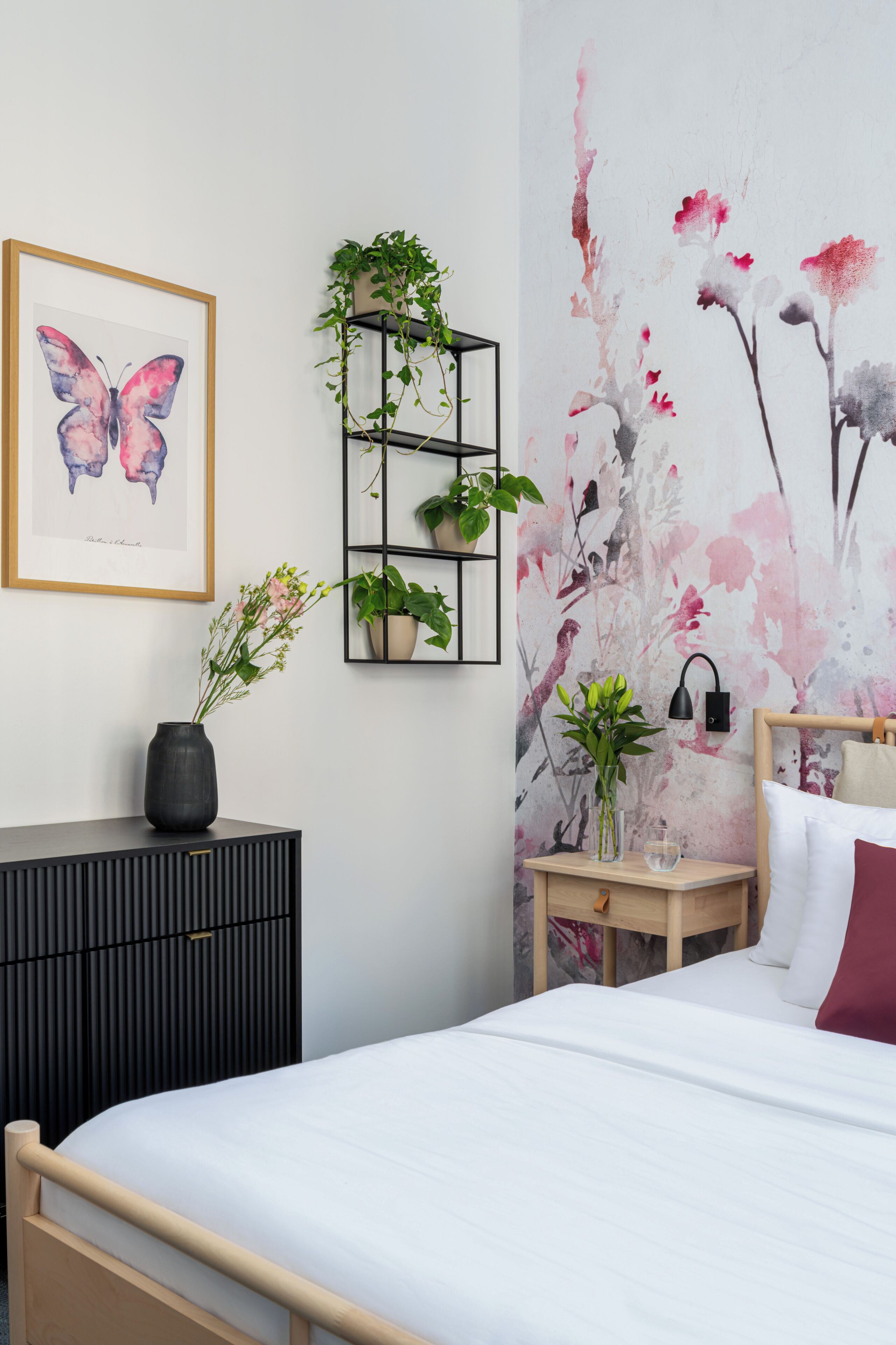 Un dormitorio con cama, mesita de noche, estante negro con plantas, una obra de arte con mariposas enmarcada y papel tapiz floral.