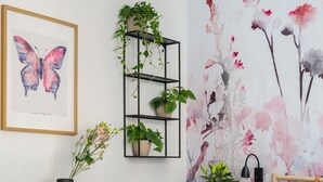 Une chambre à coucher comprenant un lit, une table de chevet, une commode, une étagère noire ornée de plantes, une œuvre d’art représentant un papillon encadré et un papier peint à motifs floraux.
