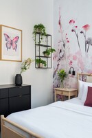 Un dormitorio con cama, mesita de noche, estante negro con plantas, una obra de arte con mariposas enmarcada y papel tapiz floral.