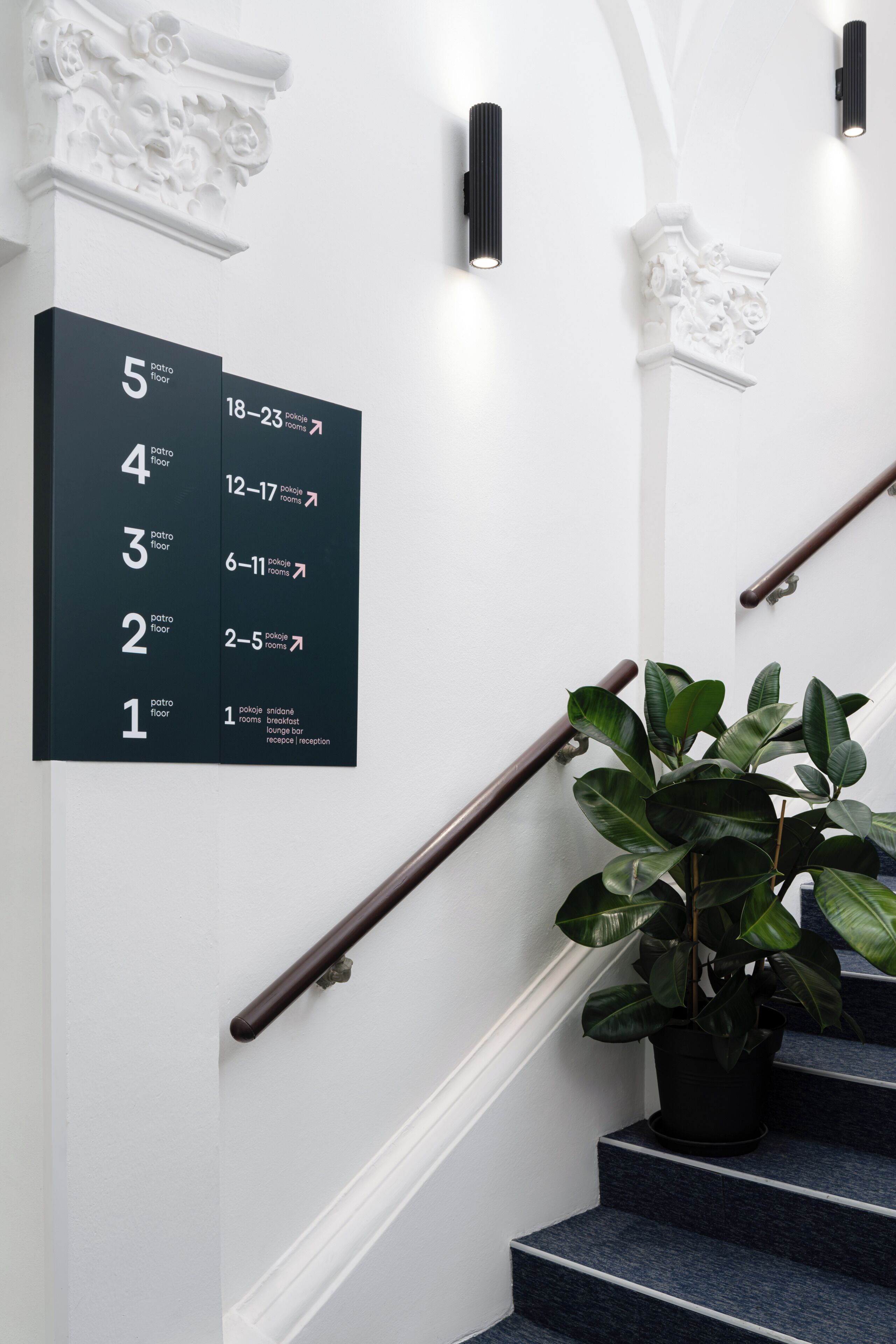 A staircase with a black railing, a potted plant, and a wall-mounted sign indicating room numbers and corresponding room types.