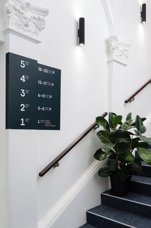 Un escalier avec une rampe noire, un pot de fleurs et un panneau fixé au mur indiquant les numéros des chambres et les types de chambres correspondants.