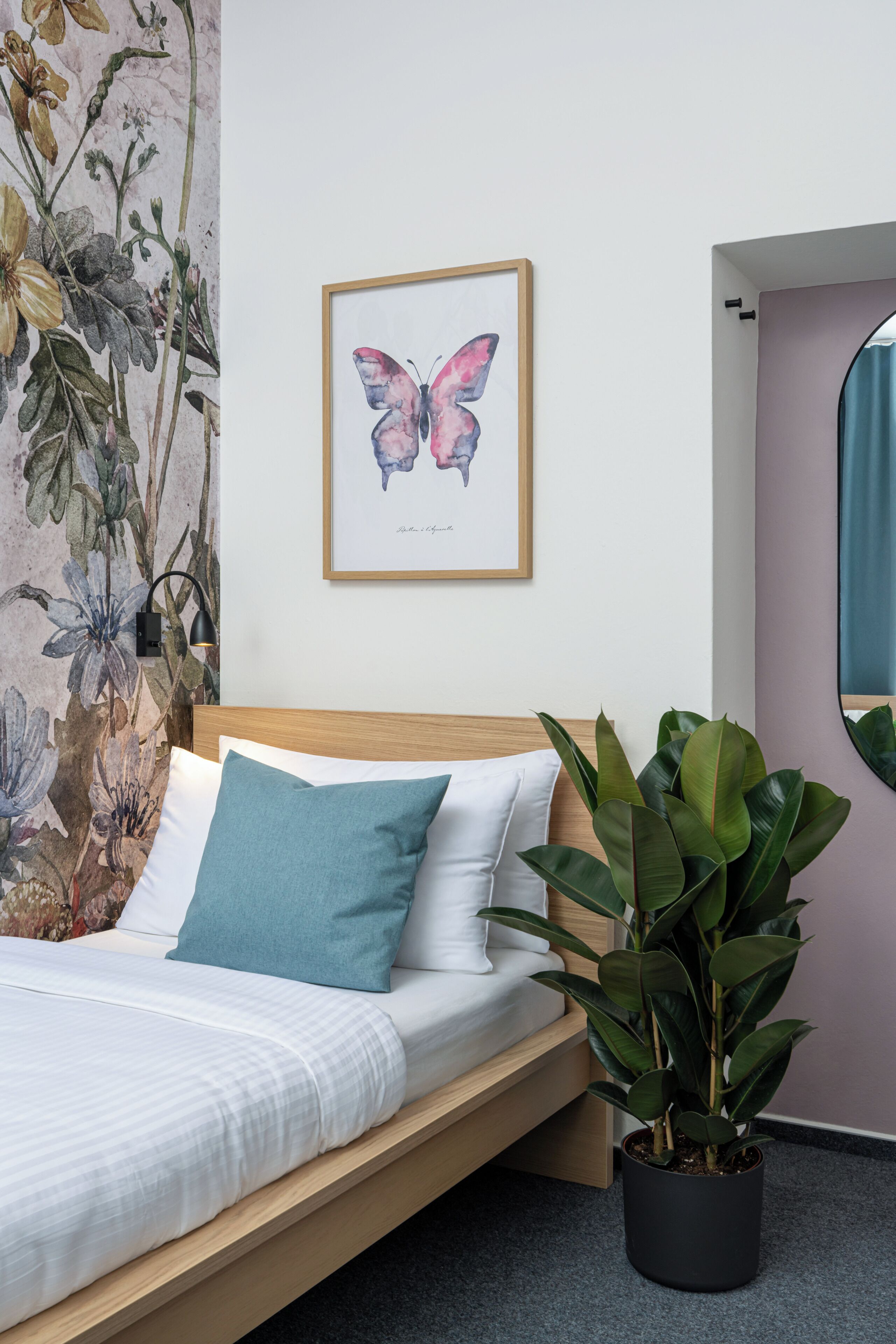 Ein Bett mit Holz-Kopfteil, einem blauen Kissen und einem weißen Kissen. An der Wand hängt ein gerahmtes Schmetterlingsbild.